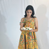 Woman in a colorful patterned dress holding a plate of food against a white curtain background